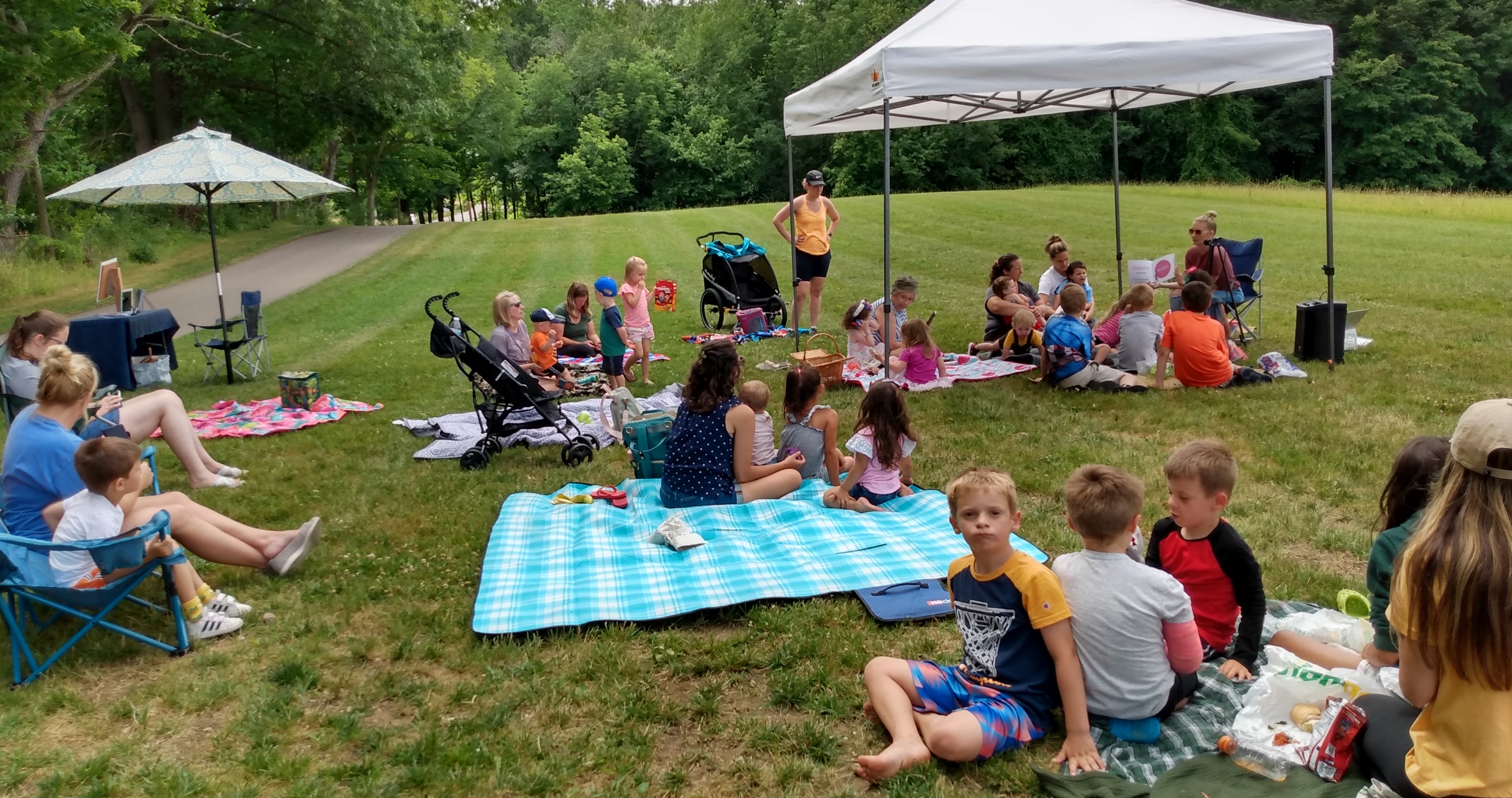 large group of people with children, seated on a grassy lawn with blankets and popup tents. One adult is reading a storybook.